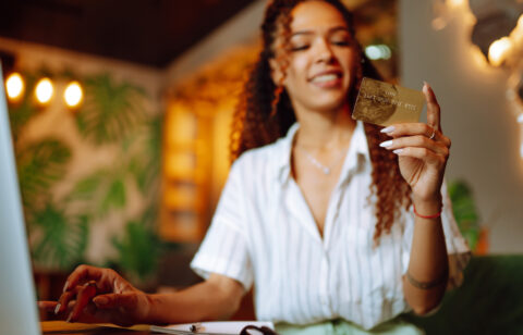 Smiling woman holding a gold credit card while sitting at a desk with a laptop.