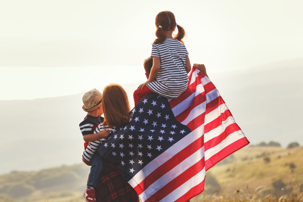 "Family standing outdoors with two children and parents holding an American flag across their backs at sunset.