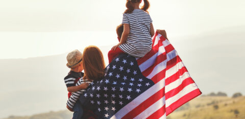 "Family standing outdoors with two children and parents holding an American flag across their backs at sunset.