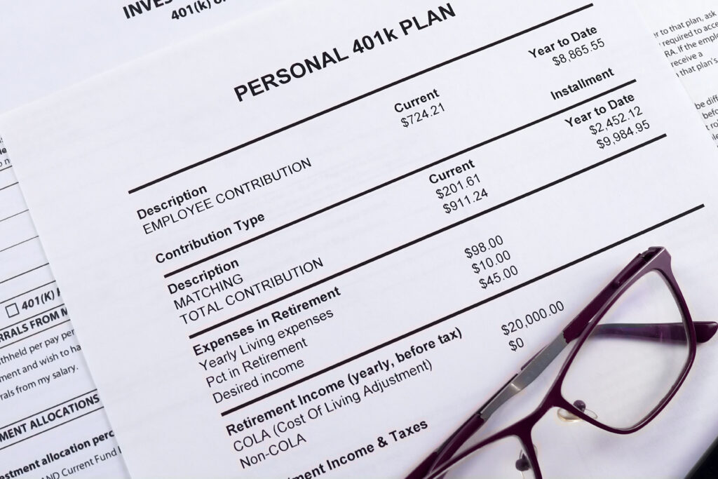 Close-up of a personal 401k plan statement showing employee contributions, matching amounts, and retirement income details, with a pair of eyeglasses resting on the paper.
