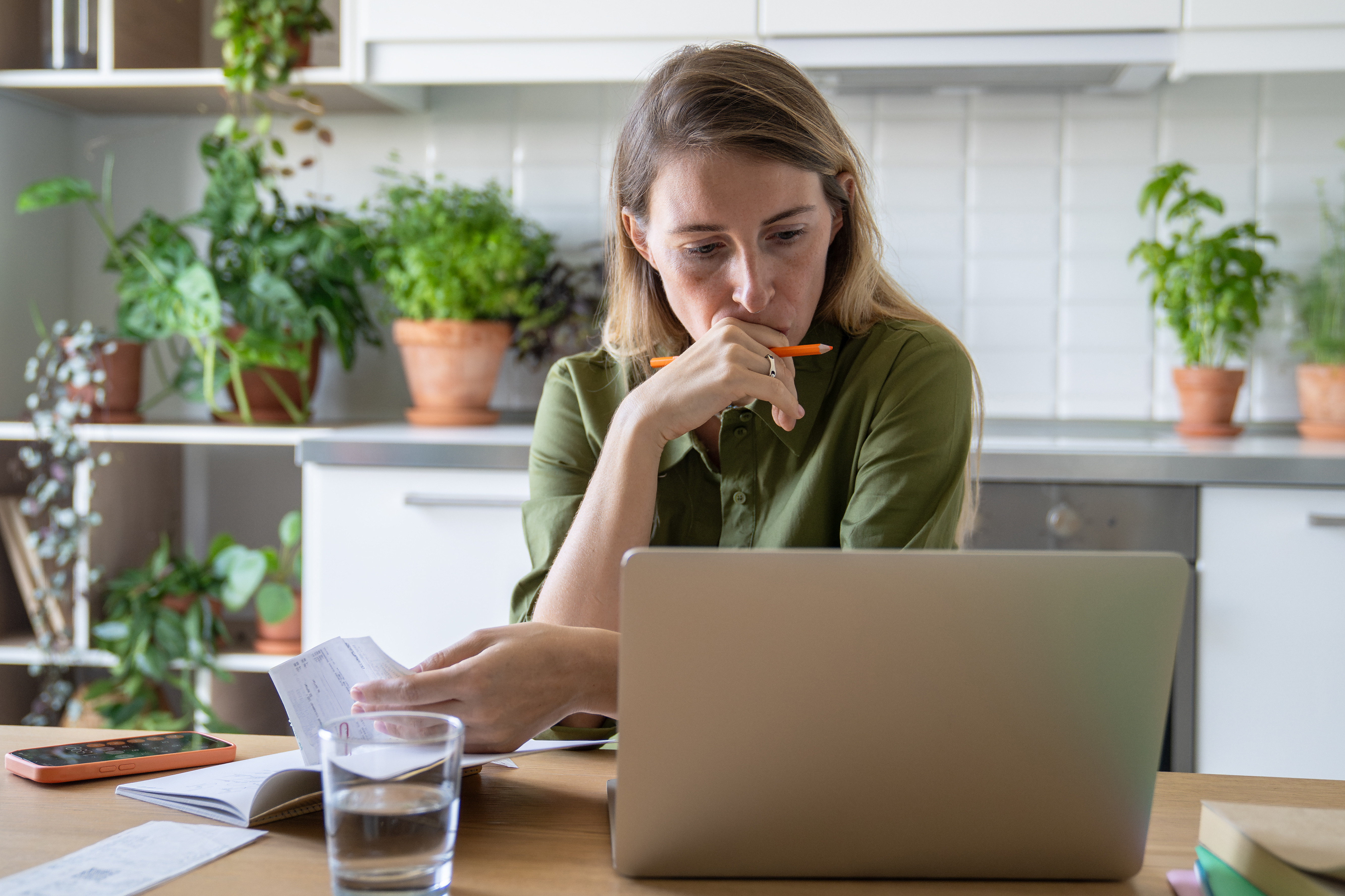 Woman sitting at a kitchen table, reviewing bills and using a laptop with a thoughtful expression.