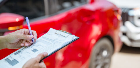Person filling out an insurance claim form on a clipboard near a red car after an accident