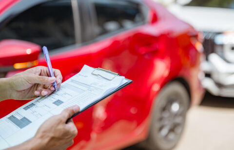 Person filling out an insurance claim form on a clipboard near a red car after an accident