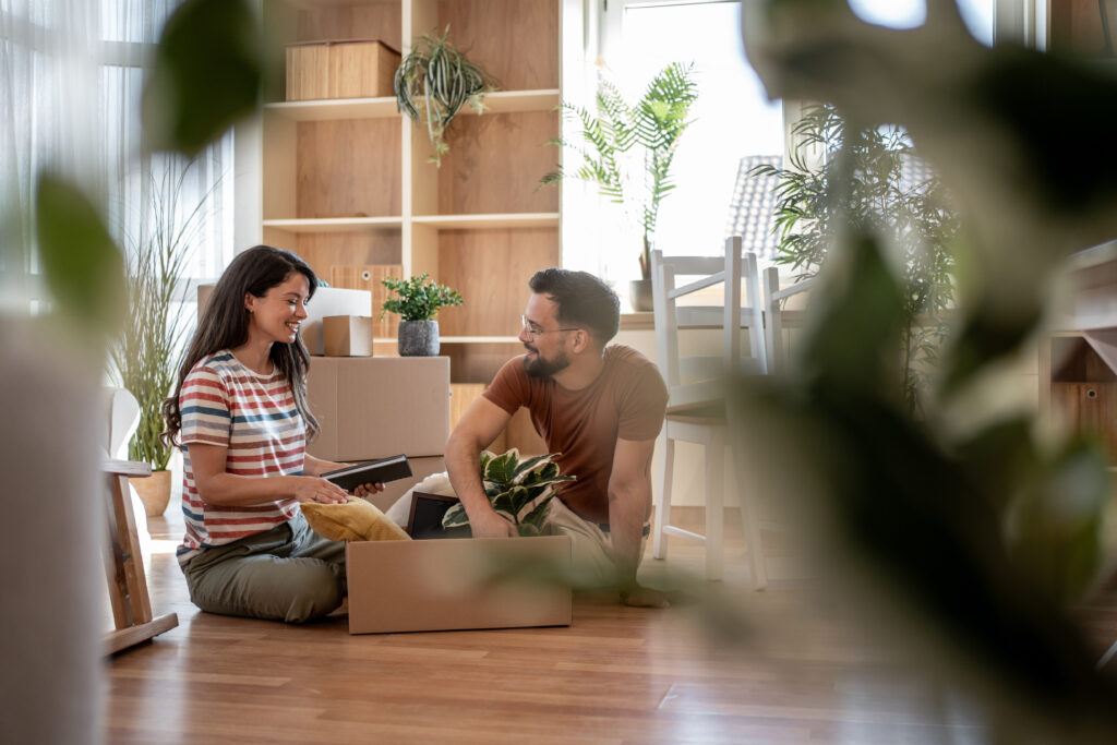 Smiling couple sits on the floor unpacking boxes and decorating their new home with plants.