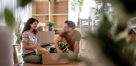 Smiling couple sits on the floor unpacking boxes and decorating their new home with plants.