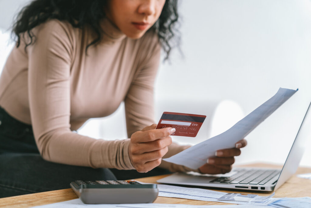 Woman reviewing a bill while holding a credit card, with a calculator and laptop on a desk.