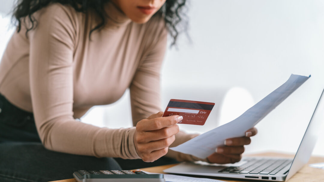 Woman reviewing a bill while holding a credit card, with a calculator and laptop on a desk.
