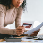 Woman reviewing a bill while holding a credit card, with a calculator and laptop on a desk.