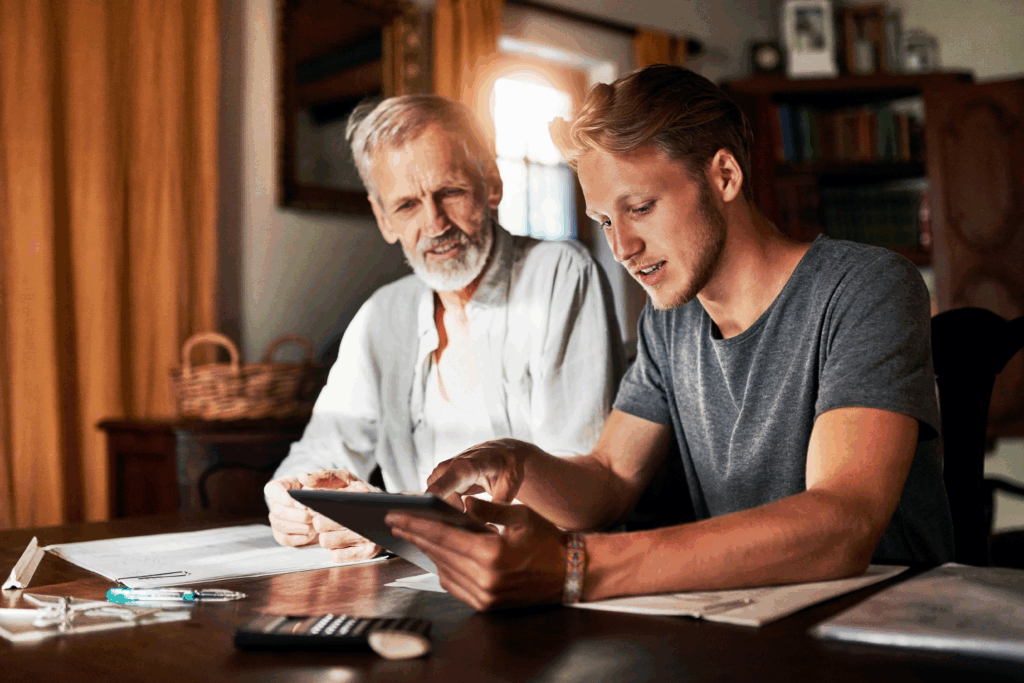 A young man showing an older man something on a tablet while they sit together at a table covered with papers and a calculator.