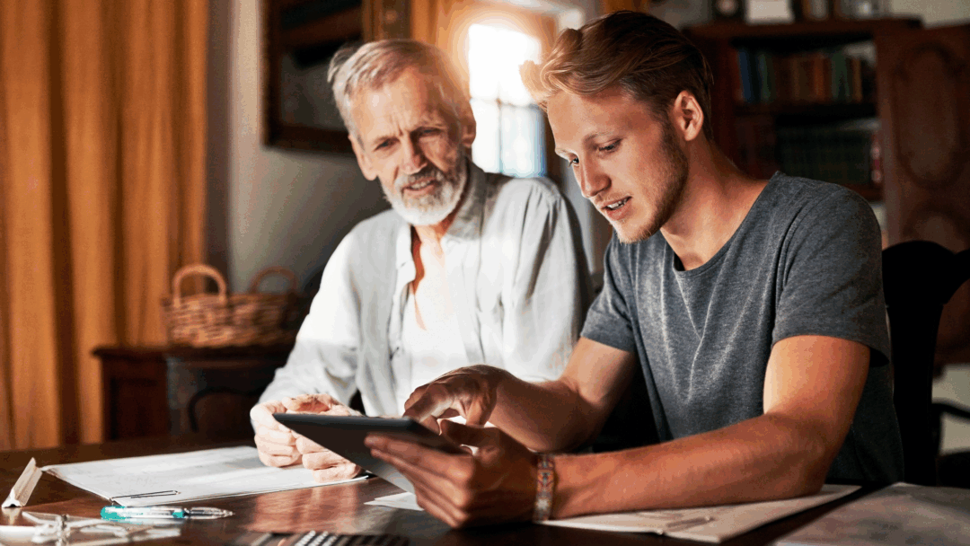 A young man showing an older man something on a tablet while they sit together at a table covered with papers and a calculator.