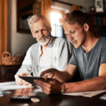 A young man showing an older man something on a tablet while they sit together at a table covered with papers and a calculator.