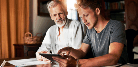 A young man showing an older man something on a tablet while they sit together at a table covered with papers and a calculator.