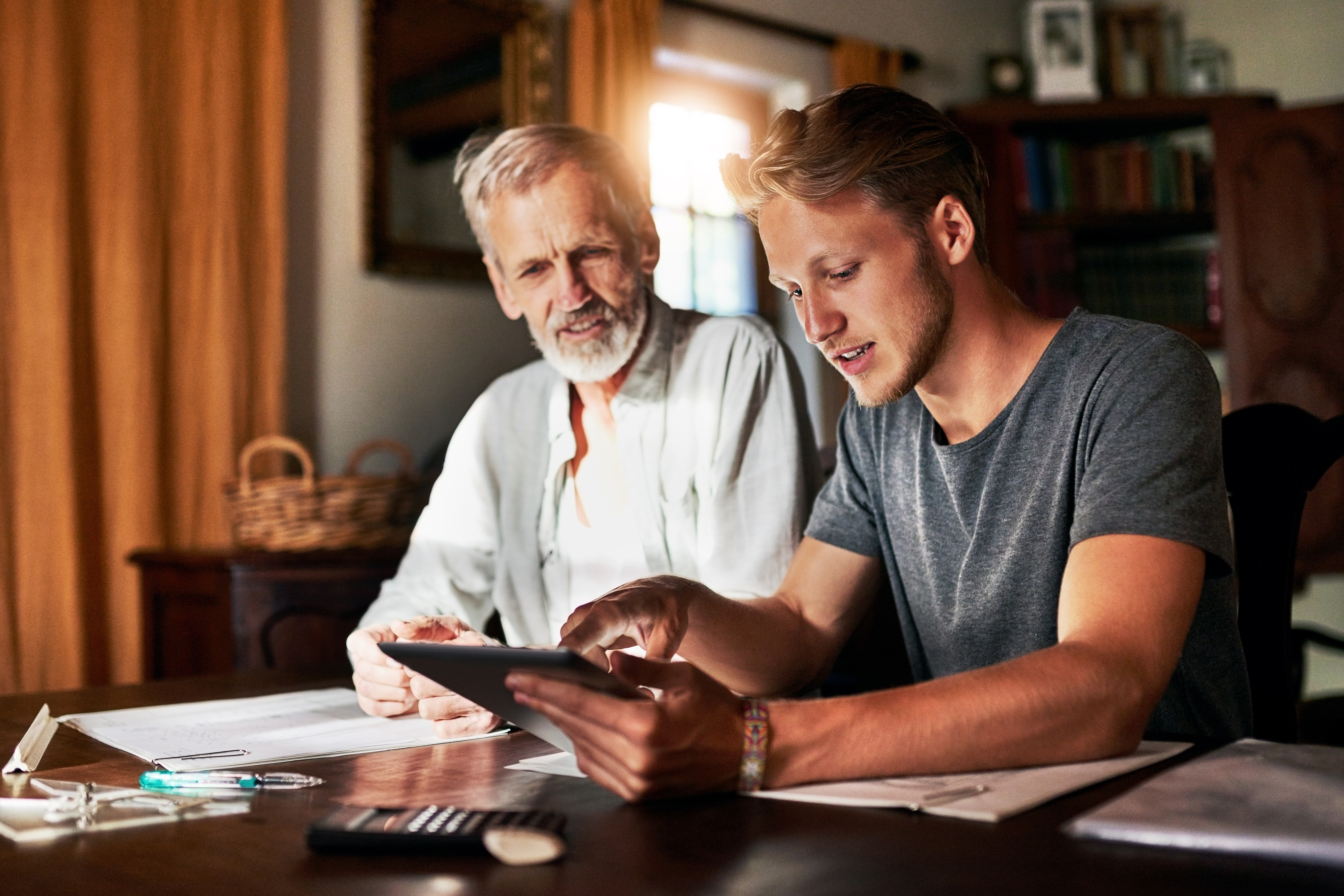 A young man showing an older man something on a tablet while they sit together at a table covered with papers and a calculator.