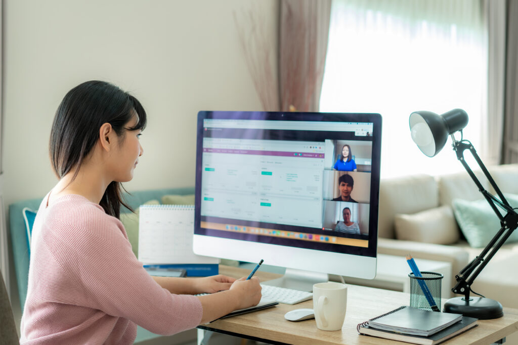 Woman working from home attends an online meeting on her computer while taking notes at her desk.