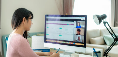 Woman working from home attends an online meeting on her computer while taking notes at her desk.