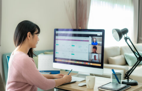 Woman working from home attends an online meeting on her computer while taking notes at her desk.