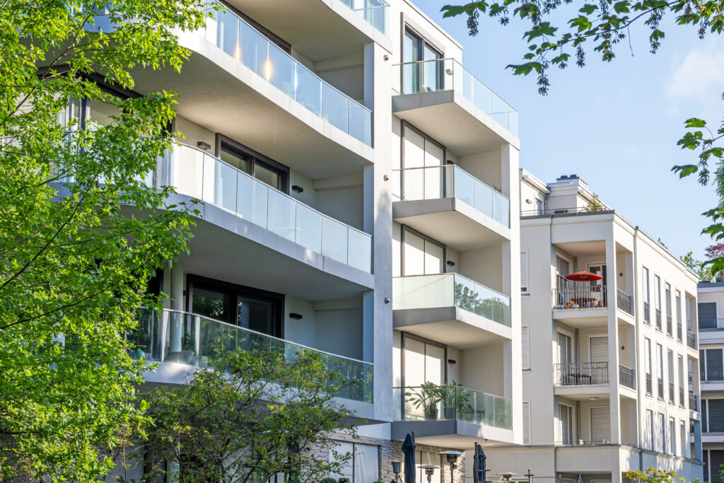 Modern apartment building with glass balconies surrounded by green trees on a sunny day.