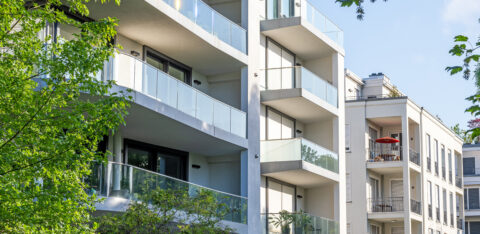 Modern apartment building with glass balconies surrounded by green trees on a sunny day.