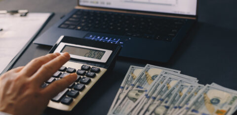 Hand pressing calculator buttons next to cash and a computer displaying a financial spreadsheet.