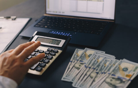 Hand pressing calculator buttons next to cash and a computer displaying a financial spreadsheet.