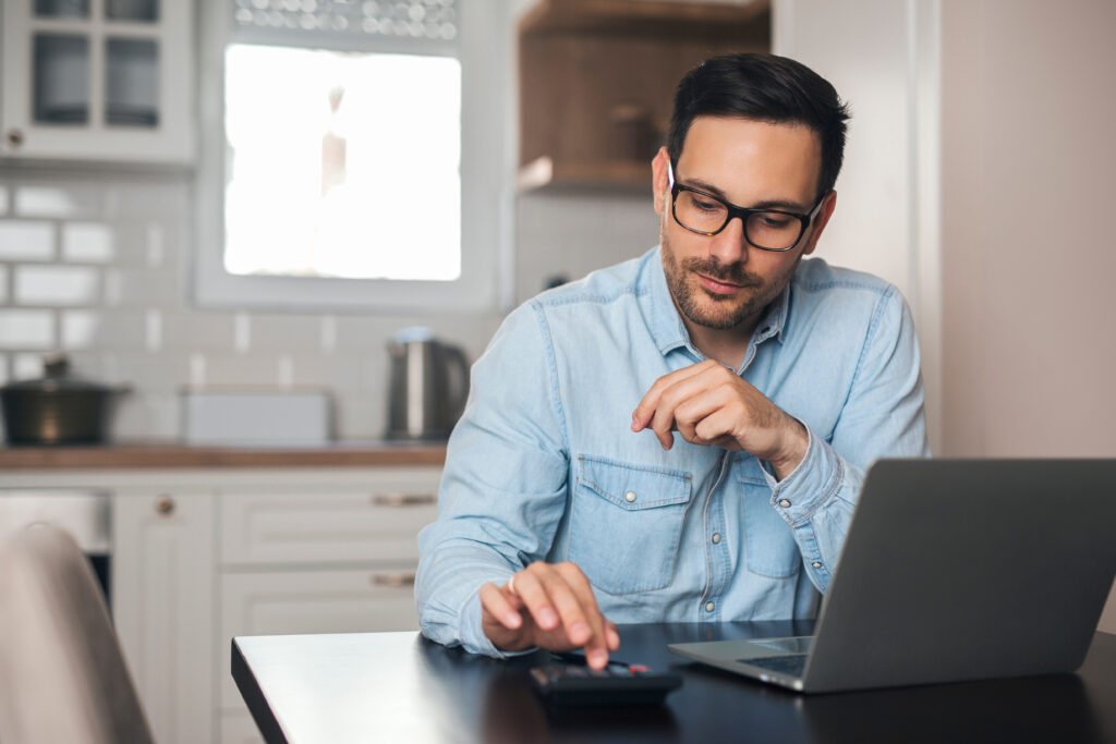 A man using a calculator while working on a laptop at his kitchen table.