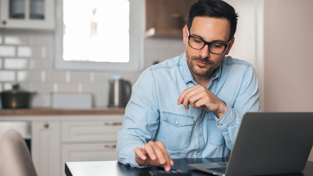 A man using a calculator while working on a laptop at his kitchen table.