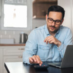 A man using a calculator while working on a laptop at his kitchen table.