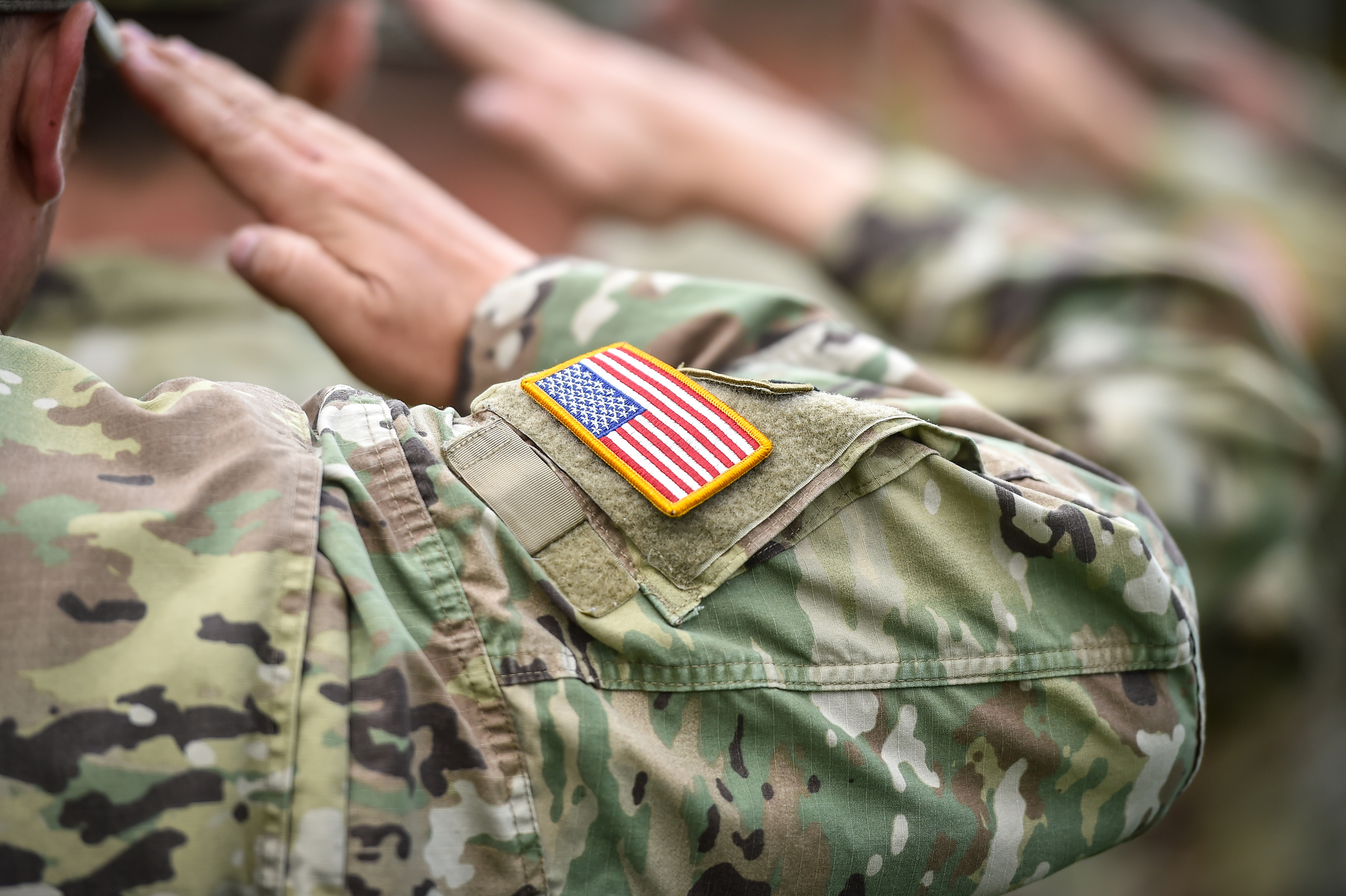 U.S. service members in camouflage uniforms saluting, with an American flag patch visible on one sleeve.