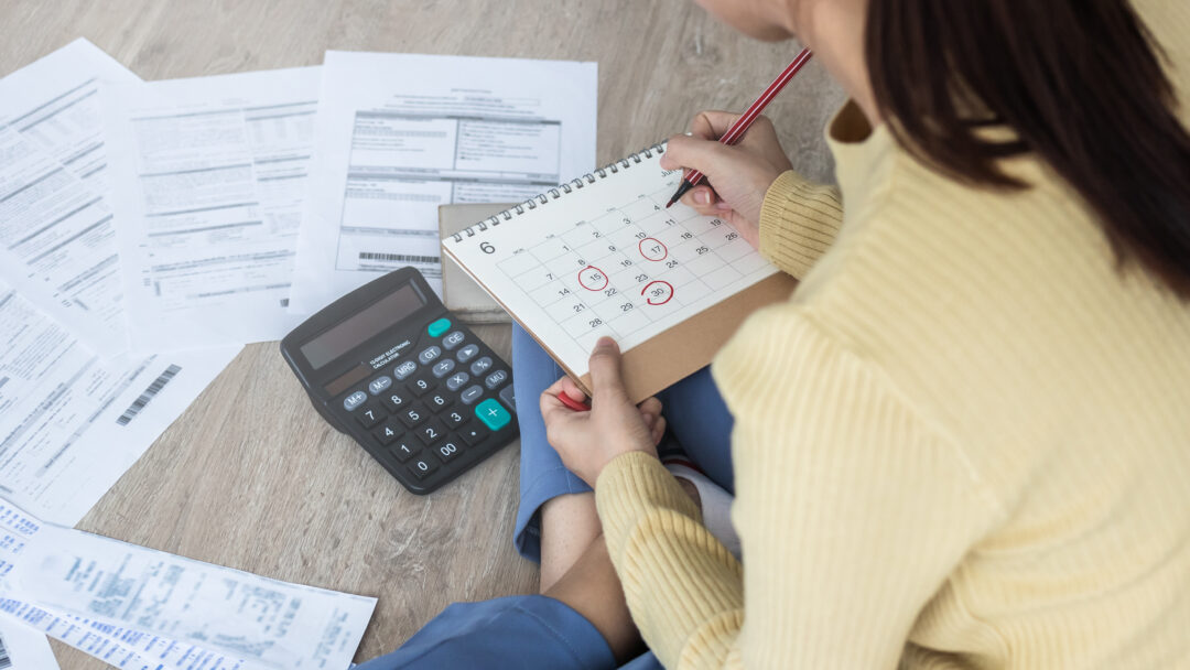 Person sitting on the floor with bills, a calculator, and a calendar, marking due dates for payments.