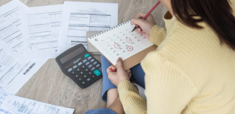 Person sitting on the floor with bills, a calculator, and a calendar, marking due dates for payments.