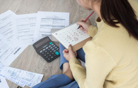 Person sitting on the floor with bills, a calculator, and a calendar, marking due dates for payments.