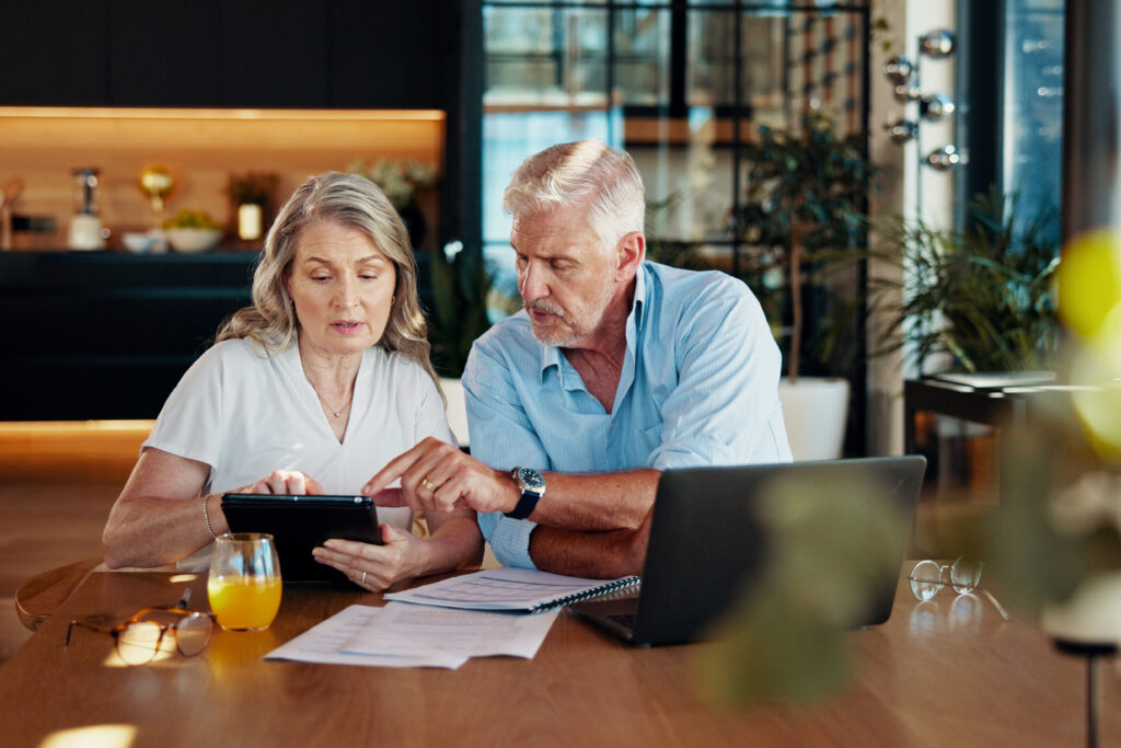 Older couple sits at a table reviewing financial documents together while looking at a tablet.