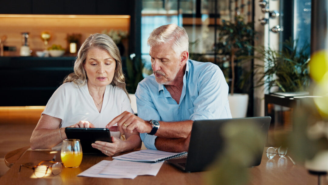 Older couple sits at a table reviewing financial documents together while looking at a tablet.