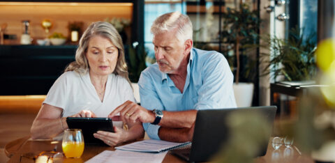 Older couple sits at a table reviewing financial documents together while looking at a tablet.