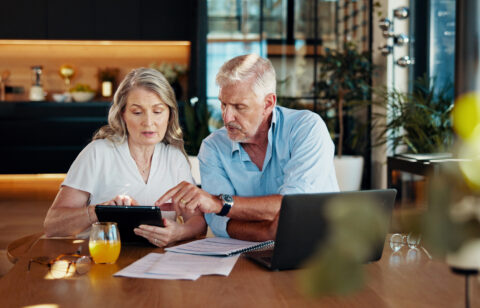 Older couple sits at a table reviewing financial documents together while looking at a tablet.