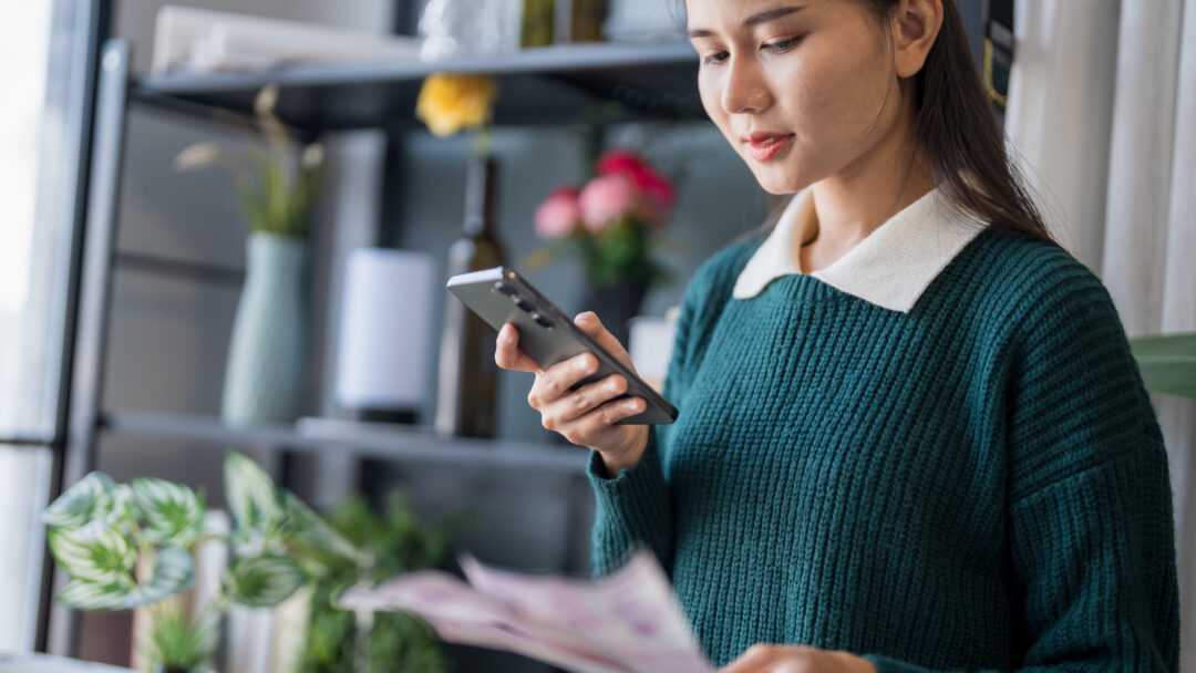 Young woman using her phone to review bills or financial documents at home.