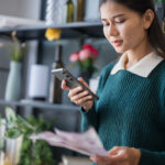 Young woman using her phone to review bills or financial documents at home.