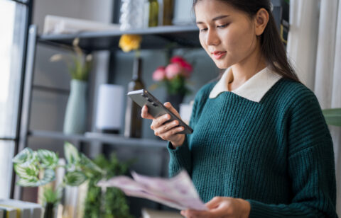 Young woman using her phone to review bills or financial documents at home.