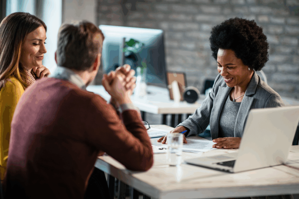 A smiling business professional meets with two clients at a modern office table, reviewing paperwork together.