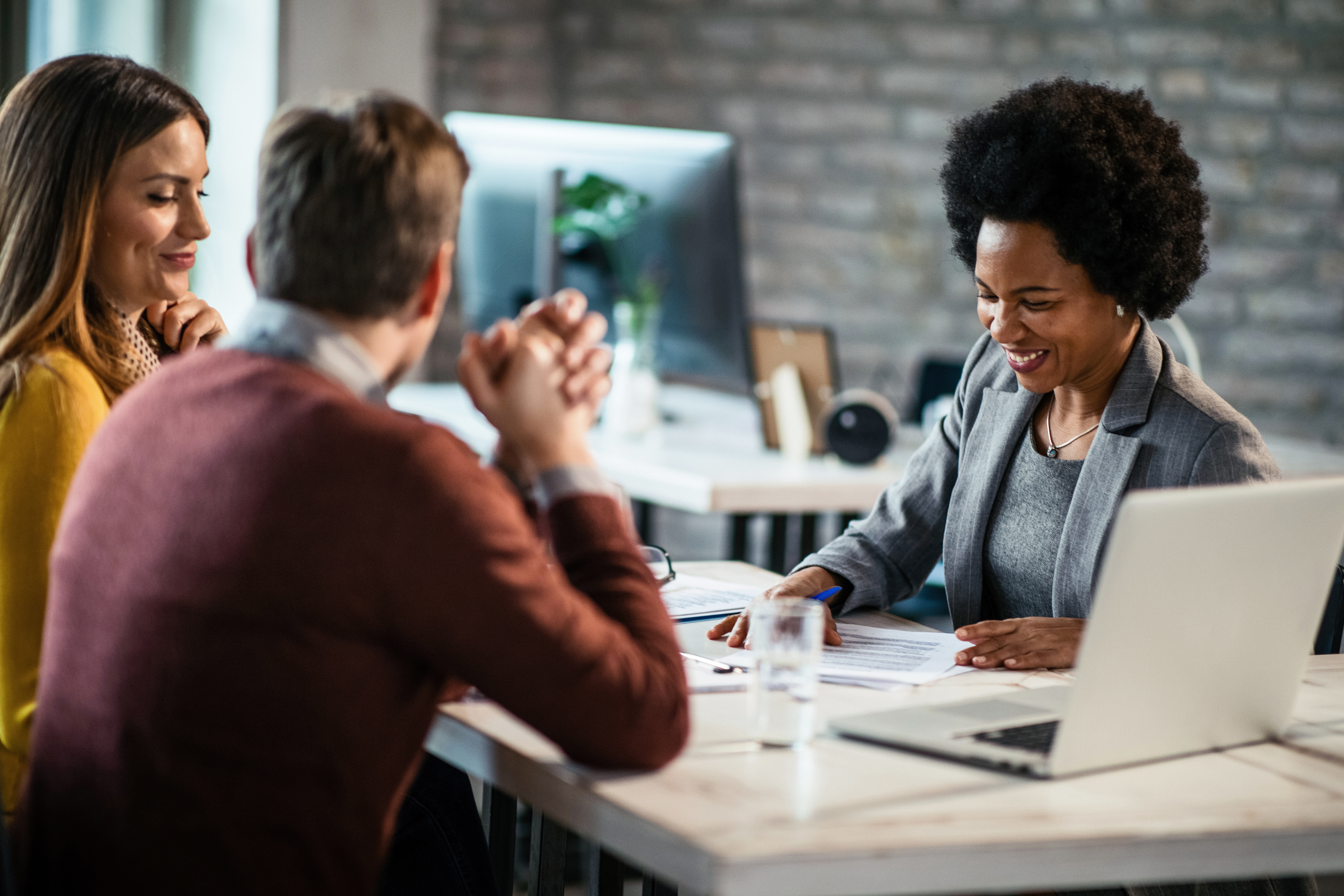 A smiling business professional meets with two clients at a modern office table, reviewing paperwork together.