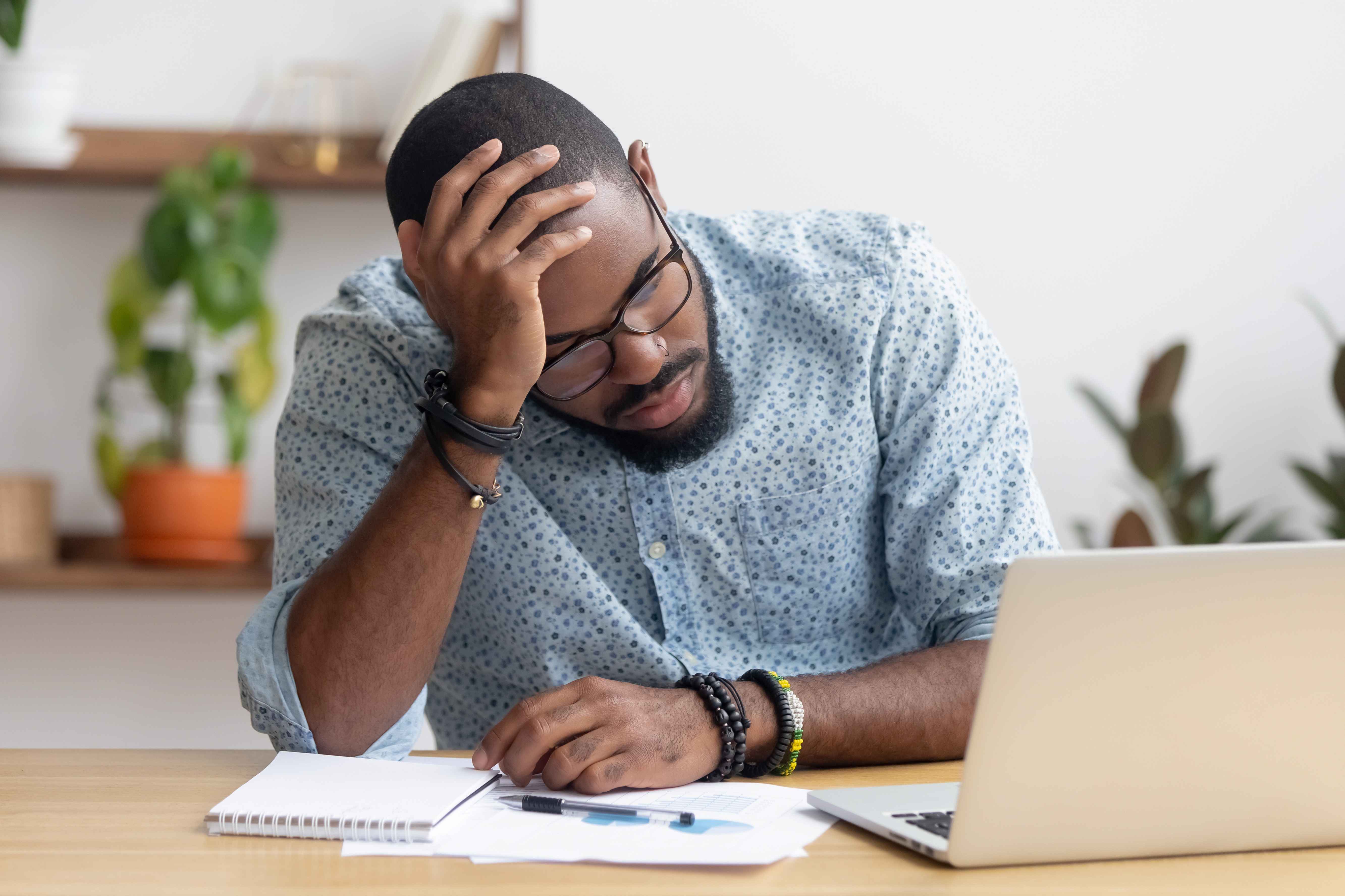 Stressed man sitting at a desk with a laptop, holding his head while reviewing financial papers.