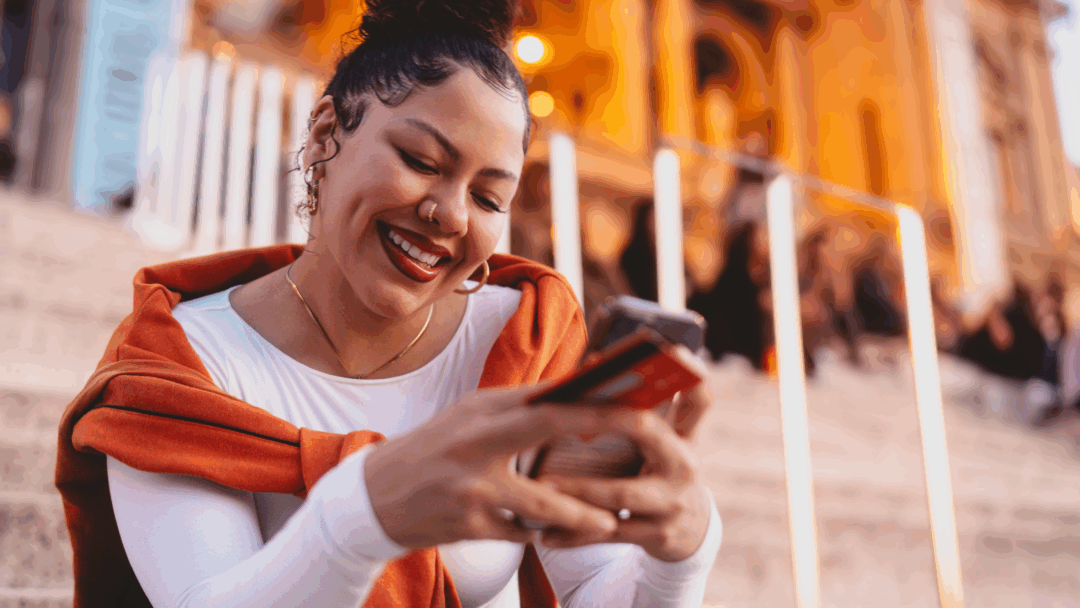 A smiling woman sits on outdoor steps in the evening, holding a phone and credit card while looking at her screen.