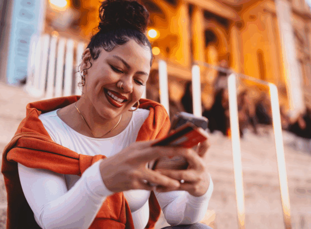 A smiling woman sits on outdoor steps in the evening, holding a phone and credit card while looking at her screen.