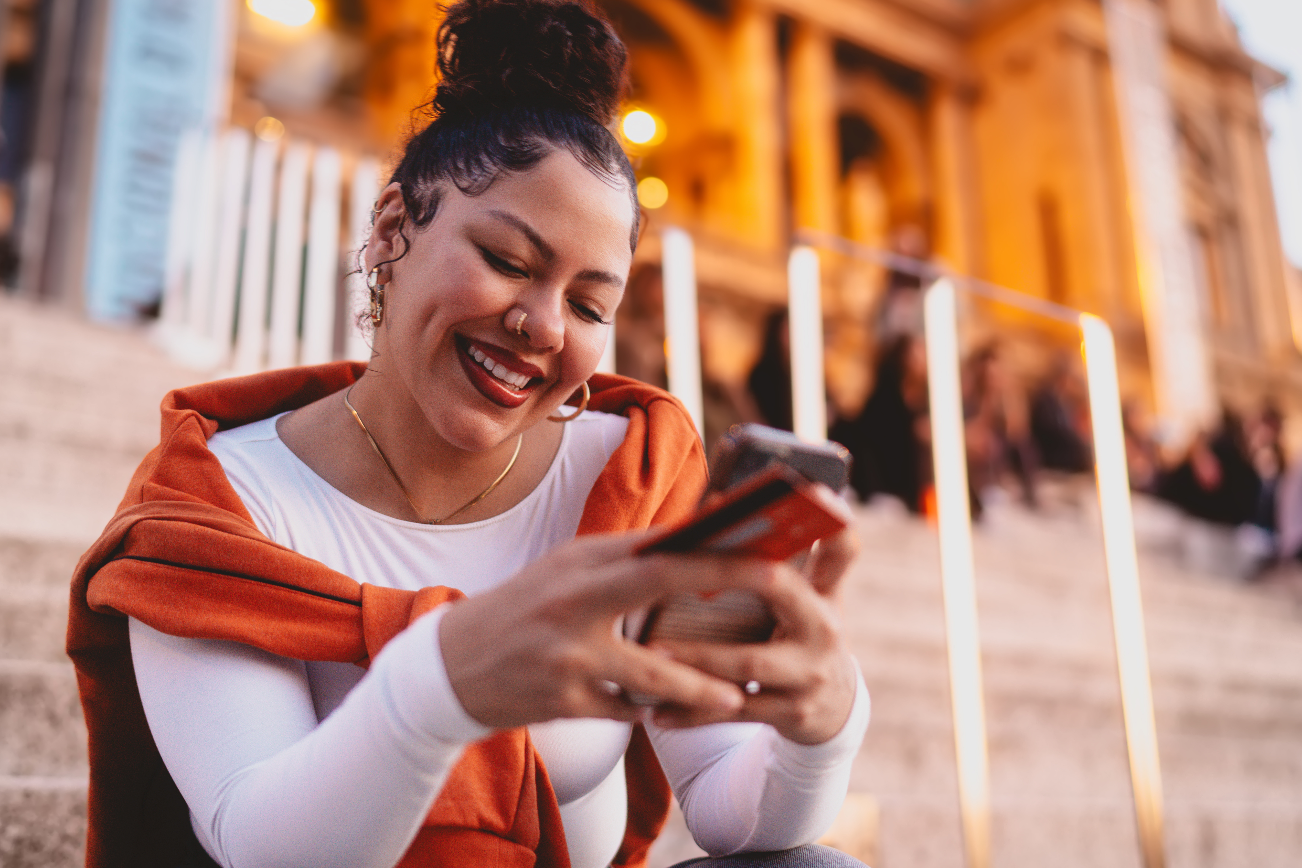 A smiling woman sits on outdoor steps in the evening, holding a phone and credit card while looking at her screen.