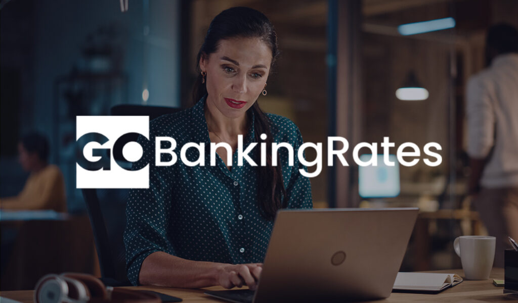 A woman works on her laptop in a dimly lit office, with the GoBankingRates logo displayed across the image.
