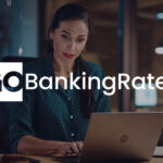 A woman works on her laptop in a dimly lit office, with the GoBankingRates logo displayed across the image.