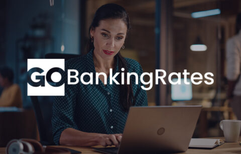 A woman works on her laptop in a dimly lit office, with the GoBankingRates logo displayed across the image.