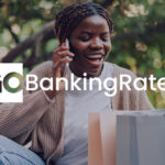 A smiling woman talks on the phone while sitting outdoors with shopping bags beside her. The GoBankingRates logo is displayed across the image.