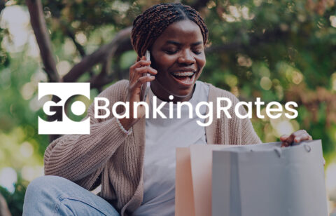 A smiling woman talks on the phone while sitting outdoors with shopping bags beside her. The GoBankingRates logo is displayed across the image.