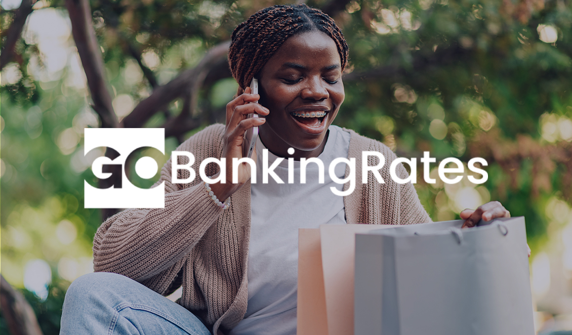 A smiling woman talks on the phone while sitting outdoors with shopping bags beside her. The GoBankingRates logo is displayed across the image.